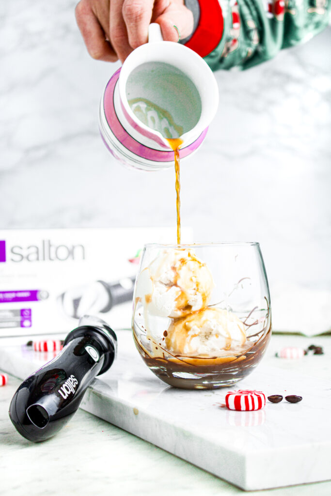 Head on view of a hand pouring a small pitcher of espresso over vegan vanilla ice cream next to an ice cream scoop and peppermint candies