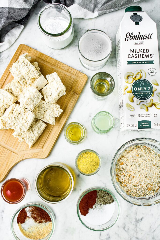 Overhead shot of all the ingredients you need to make vegan buffalo tofu wings: tofu, hot sauce, olive oil, vegan bouillon, breadcrumbs, unsweetened cashew milk, apple cider vinegar, lime juice, tapioca starch, salt, pepper, chili powder, and agave.