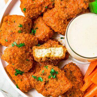 Overhead shot of a pile of breaded and sauced vegan buffalo tofu wings with a bite taken out of the center wing to show the meaty texture inside.