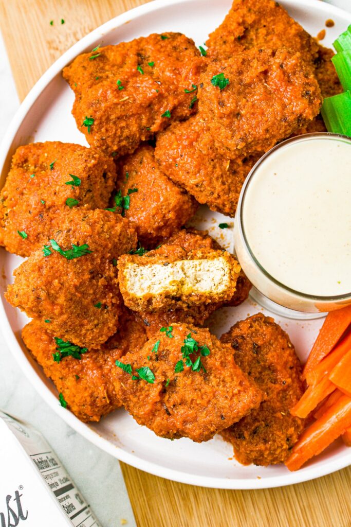 Overhead shot of a pile of breaded and sauced vegan buffalo tofu wings with a bite taken out of the center wing to show the meaty texture inside.