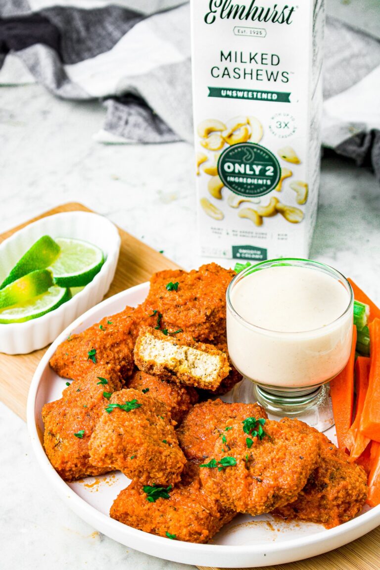 Overhead shot of a pile of vegan buffalo wings on a plate sprinkled with fresh minced parsley. One of the wings has a bite taken out of it. In the center of the plate is a glass cut holding vegan blue cheese dip, and behind the plate sits a carton of Elmhurst unsweetened cashew milk