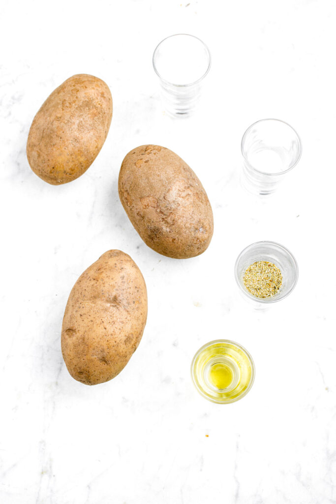 Overhead view of three potatoes, a shot glass full of oil, and another shotglass full of spices