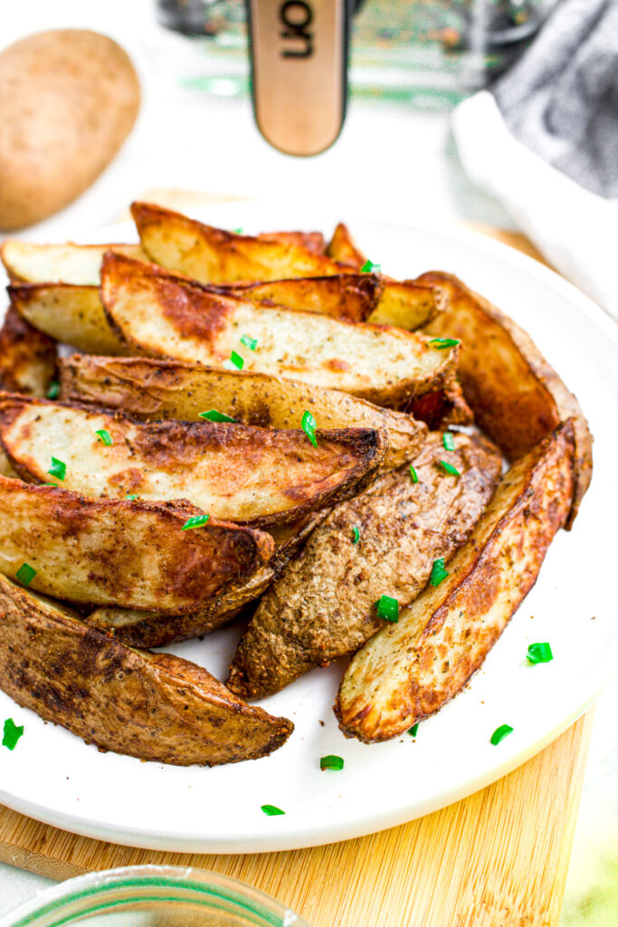 Overhead view of a pile of potato wedges on a plate in front of a clear glass air fryer basket