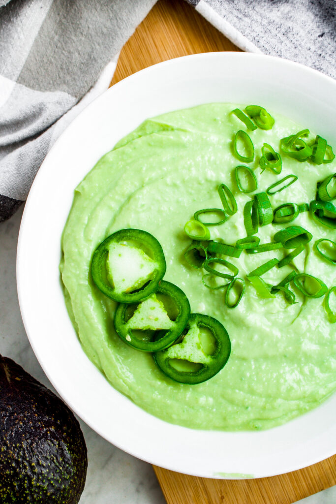 Overhead photo of a bowl of creamy avocado ranch dip topped with 3 jalapeno slices on the left and a smattering of sliced green onions on the right. A black and white checkered linen surrounds the bowl.