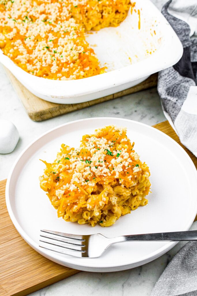A square of vegan mac and cheese on a plate with a fork next to it and a tray of more mac and cheese behind it