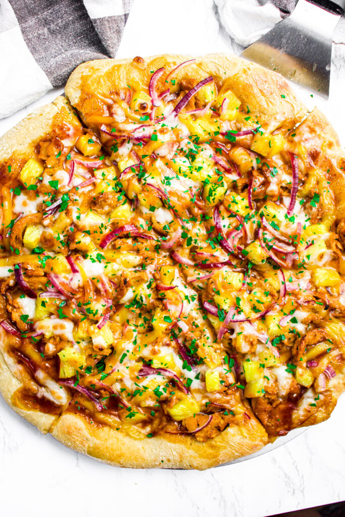 Overhead close up view of a sliced BBQ vegan pizza topped with green herbs next to a black and white checkered linen