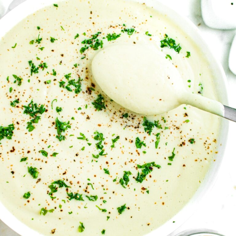 Close up overhead view of a round white bowl of easy cauliflower soup topped with parsley and pepper with a spoon pulling creamy soup out of the bowl in the top right corner