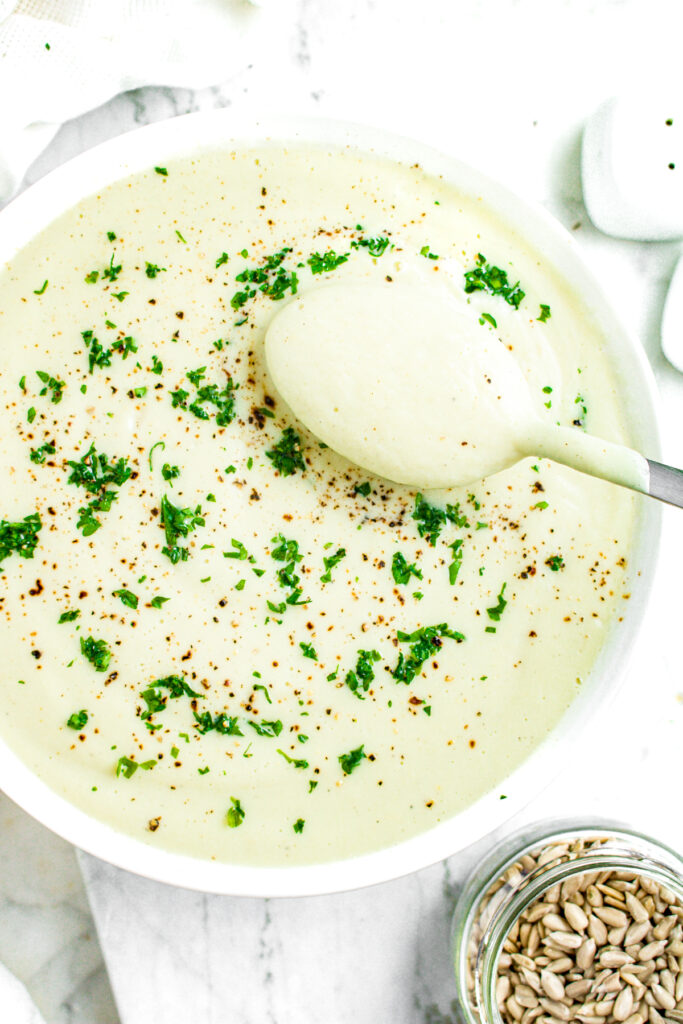 Overhead view of a round white bowl of creamy vegan cauliflower soup with a spoon pulling out of it and sunflower seeds scattered in the bottom right corner