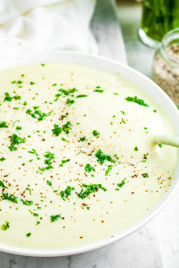 Head on view of half a round white bowl filled with cauliflower lentil soup with a spoon digging into it and a jar of parsley to the back right