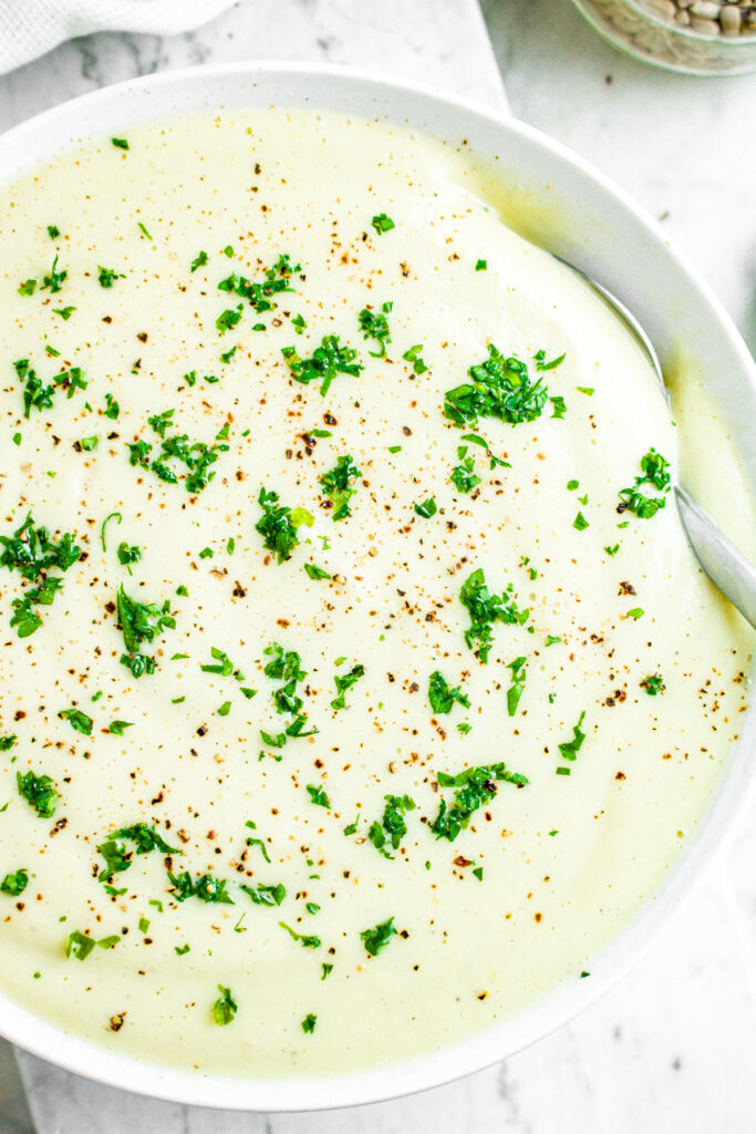 Zoomed in overhead view of a round white bowl filled with creamy dairy free cauliflower soup garnished with minced parsley and ground black pepper
