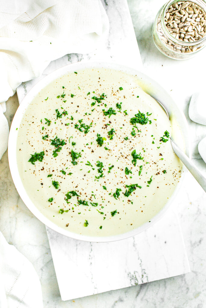 Overhead view of a round white bowl with a white linen napkin to the left and sunflower seeds in a clear glass jar to the top right and a bowl of creamy cauliflower soup in the center