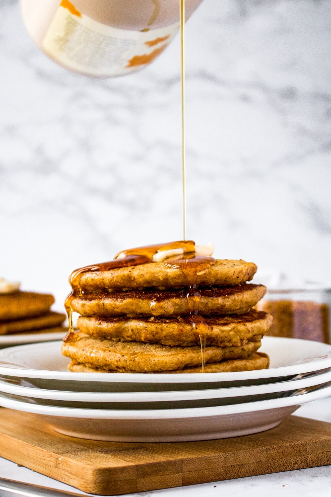 A stack of vegan pancakes with coconut bacon stacked on a small white plates topped with a square of vegan butter and a drizzle of maple syrup.