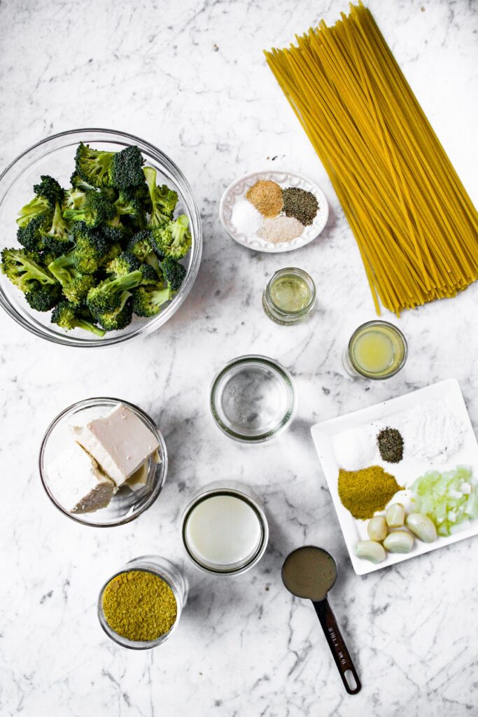 Overhead shot of the ingredients you need to make vegan broccoli alfredo: fettuccine, broccoli, olive oil, unsweetened plain non-dairy milk, nutritional yeast, garlic powder, onion powder, sea salt, black pepper, lemon juice, garlic, onion, nutritional yeast, tahini, and tapioca starch