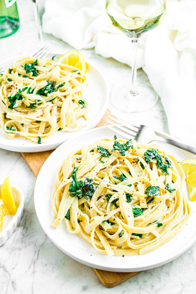 Overhead photo of two plates of creamy vegan pasta with spinach and a glass of white wine to the side