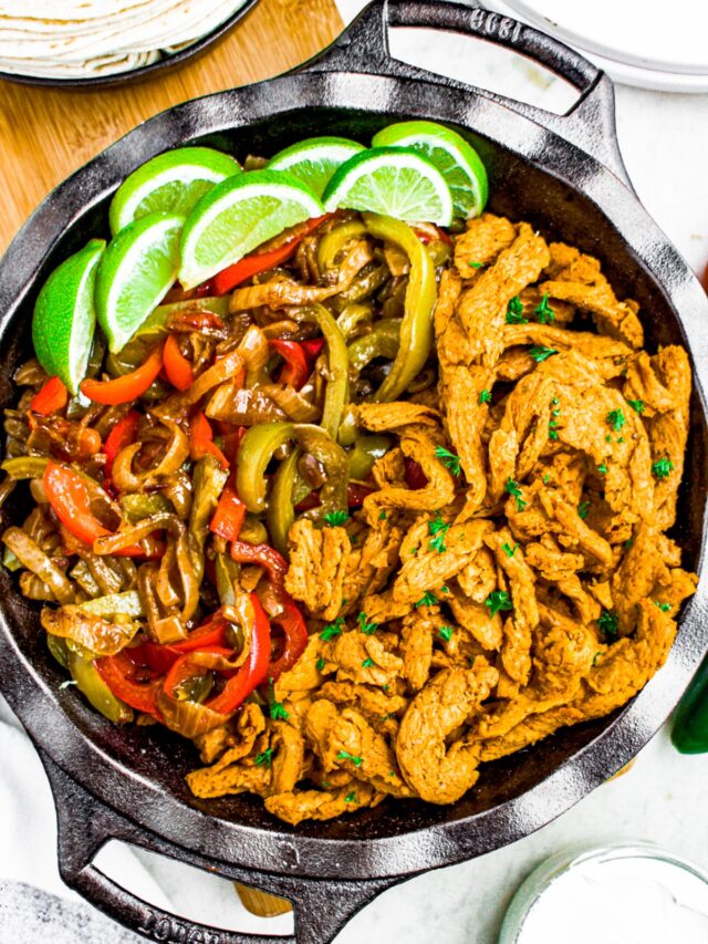 Overhead close up photo of a deep cast iron pan filled with vegan chicken, peppers and onions, and limes for fajitas