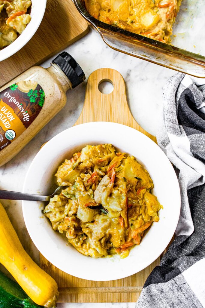 Overhead shot of a white bowl with dijon squash casserole in it surrounded by full squash and zucchini, a bottle of organicville dijon mustard, and a black and white linen