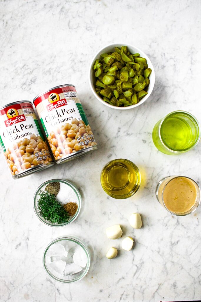 Overhead shot of the ingredients you need to make dill pickle hummus recipe: chickpeas in a can, chopped pickles, pickle juice, olive oil, cloves of garlic, tahini, salt, fresh dill, ground black pepper, ice cubes