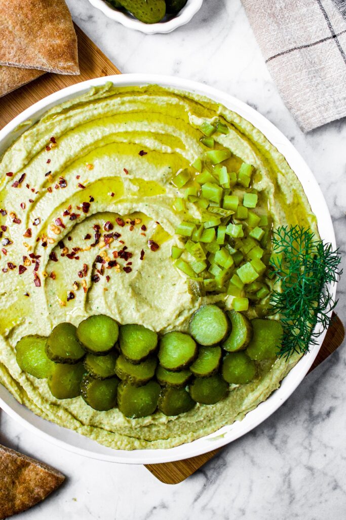 Overhead close up shot of a plate of homemade trader joes dill pickle hummus in a swirl pattern topped with sliced and chopped dill pickles, crushed red pepper flakes, and extra virgin olive oil. The plate is surrounded by air fryer pita chips.
