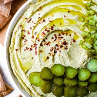 Overhead shot of a plate of homemade trader joes dill pickle hummus in a swirl pattern topped with sliced and chopped dill pickles, crushed red pepper flakes, and extra virgin olive oil. The plate is surrounded by air fryer pita chips.