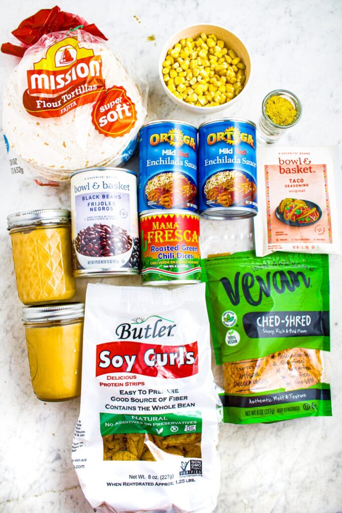 Overhead photo of all the ingredients you need to make vegan enchiladas: tortillas, soy curls, vegan cheese, corn, enchilada sauce, green chiles, taco seasoning, black beans, vegan cheese sauce, bouillon powder