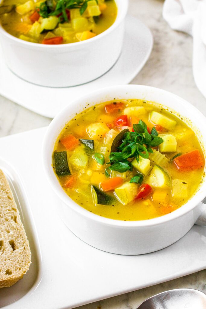 Overhead photo of two white round bowls of vegetable soup on long plates next to sliced bread