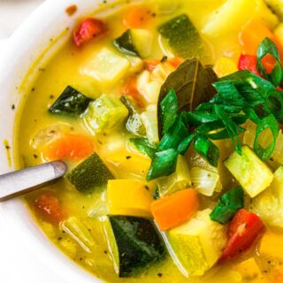Close up photo of a bowl of squash and zucchini soup with a vegan chicken broth topped with scallions in a white bowl with a spoon digging into it