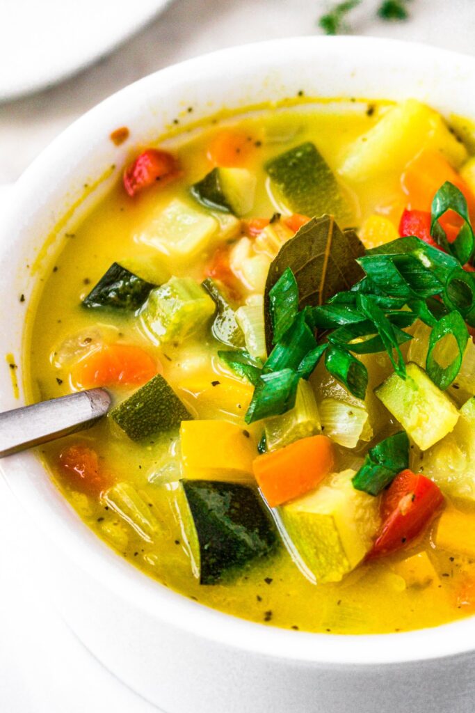 Close up photo of a bowl of squash and zucchini soup with a vegan chicken broth topped with scallions in a white bowl with a spoon digging into it