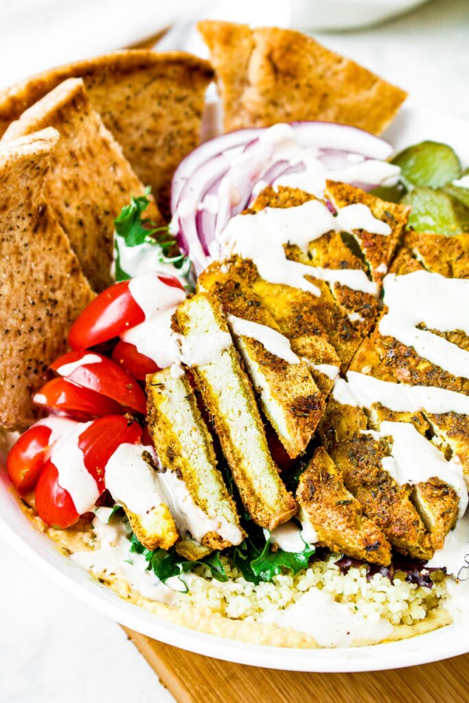 Overhead shot of a bowl filled with couscous, greens, sliced cherry tomatoes, red onions, sliced pickles, and baked Greek tofu drizzled with creamy tahini sauce