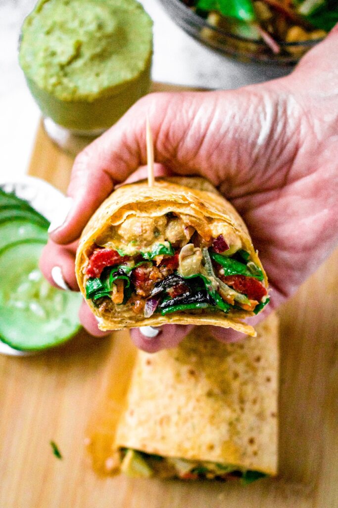 Overhead shot of a hand holding a hummus and veggie wrap that's been cut in half. There are sliced cucumbers and a dish of hummus in the background.