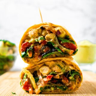 Head-on shot of a veggie wrap cut into halves and stacked on top of each other, held together with a toothpick. There is a jar of hummus and bowl of salad in the background.