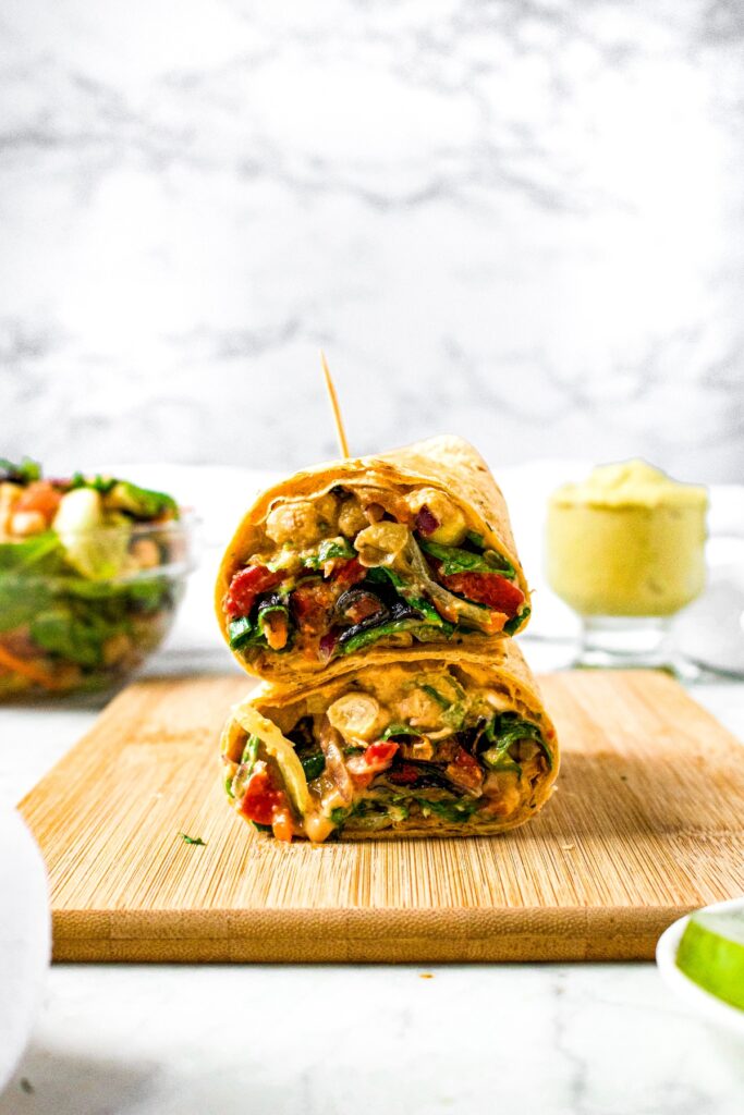 Head-on shot of a veggie wrap on a wooden board cut into halves and stacked on top of each other, held together with a toothpick. There is a jar of hummus and bowl of salad in the background.