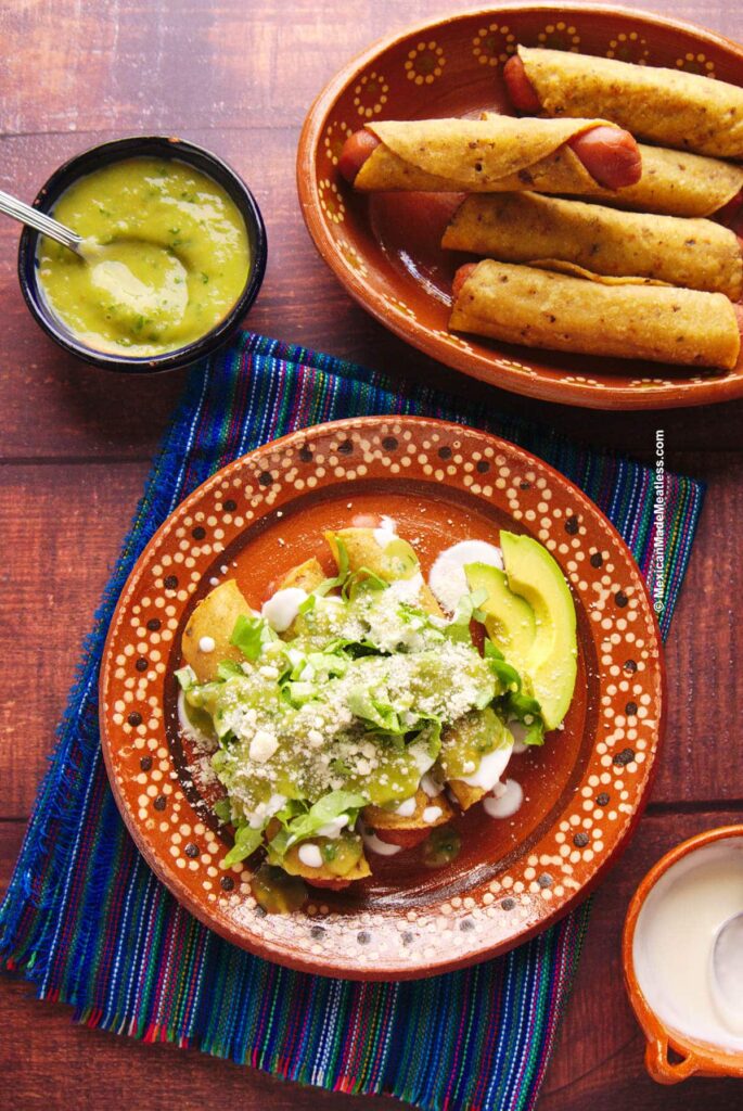 Overhead view of two plates: one with three tacos covered in shredded veggies and creamy sauce and the other filled with tortilla wrapped hot dogs