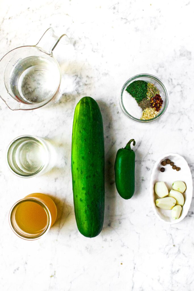 Overhead shot of the ingredients you need to make no cook refrigerator pickles: a cucumber, jalapeno, apple cider vinegar, white vinegar, hot water, garlic cloves, all spice berries, sugar, pepper, dehydrated onion, red pepper, black pepper, fennel, and fresh dill