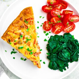 Overhead photo of a round white plate with a slice of vegan egg casserole, spinach, and sliced grape tomatoes sprinkled with minced green onion