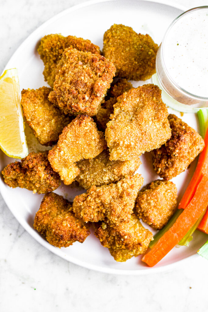 Overhead close up view of breaded tofu crispy tofu pieces on a round white plate with a lemon wedge and carrot and celery sticks