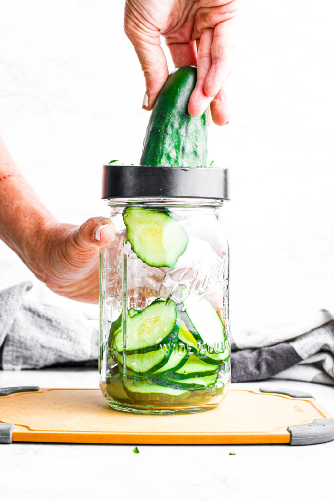 Head on view of a hand using a jar top slicer to slice cucumbers into a large mason jar