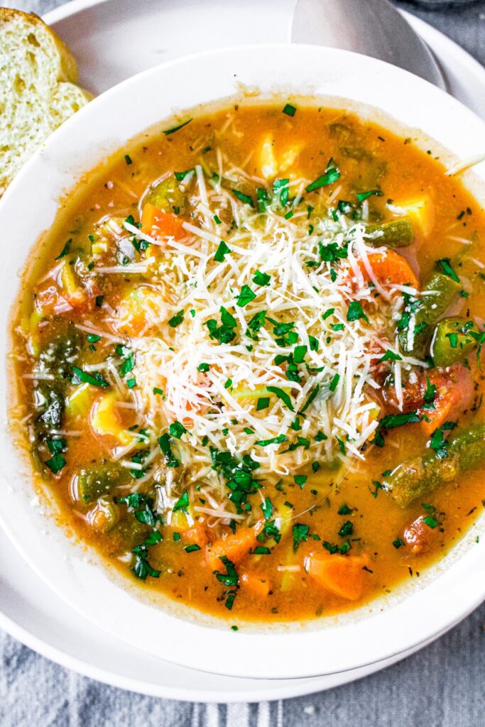 Overhead close up photo of an Italian vegetable soup in a round white bowl topped with vegan parmesan cheese
