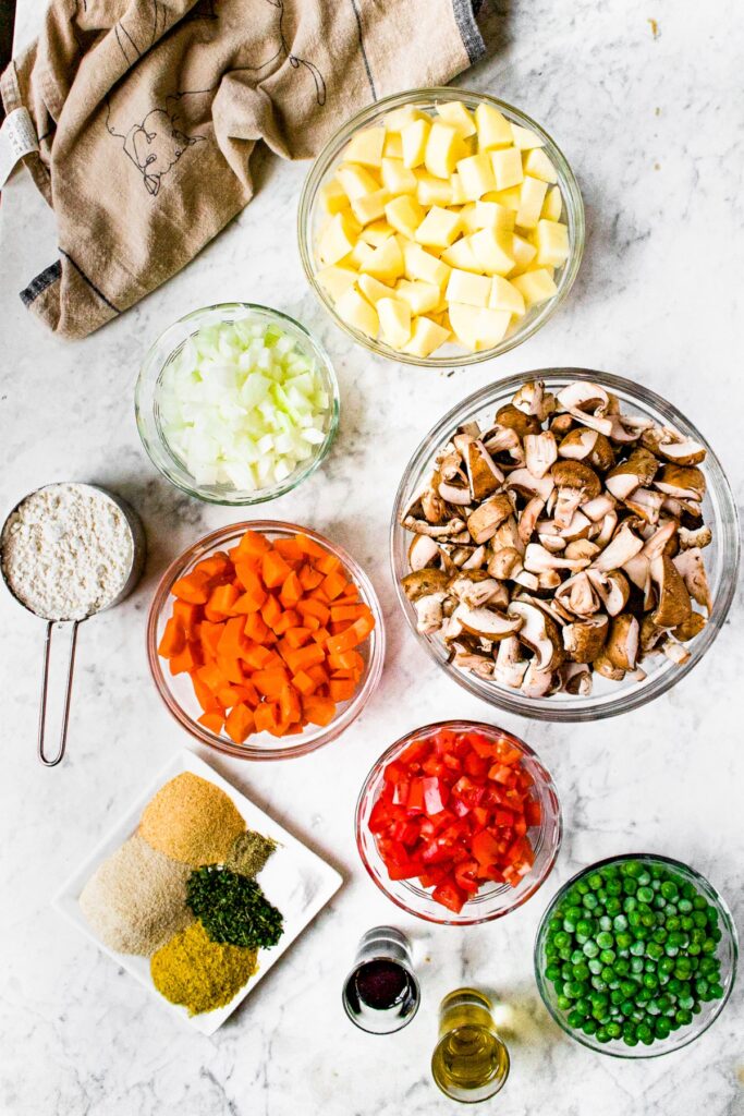Overhead shot of the ingredients you need to make vegan mushroom stew: potatoes, mushrooms, onions, carrots, all purpose flour, peas, spices, olive oil, and Gravy Master.