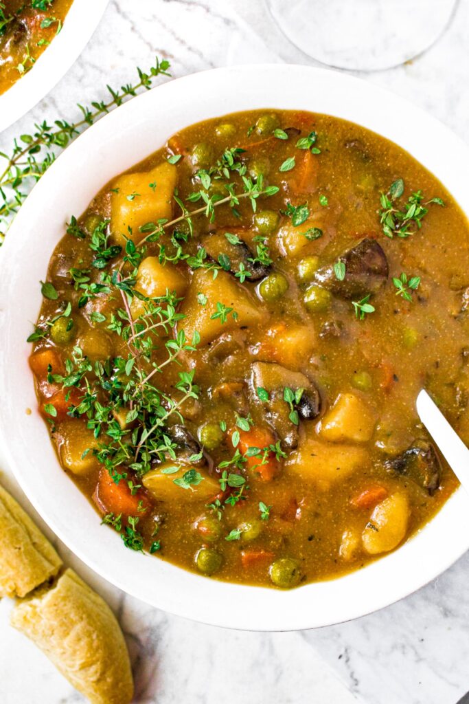 Overhead close up shot of a white bowl filled with vegan mushroom stew with a large sprig of thyme on top and a spoon digging into the stew.