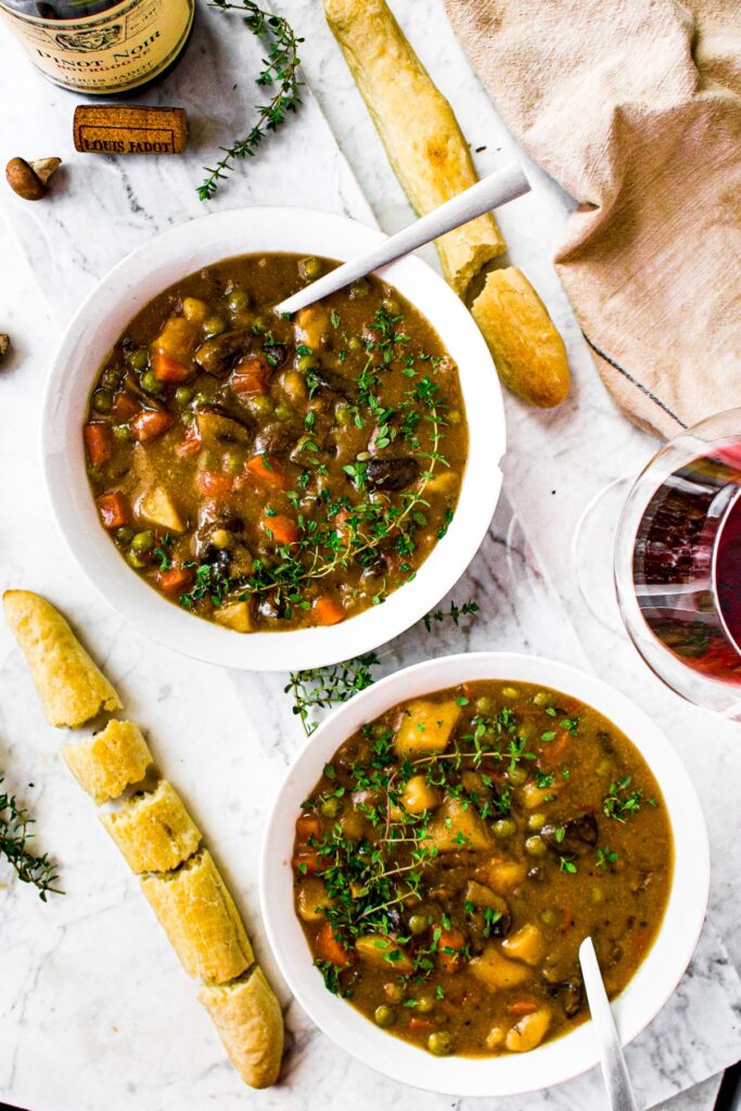 Overhead photo of two white bowls of vegetarian mushroom stew with sprigs of thyme and a spoon digging into each. There are broken up breadsticks next to each bowl and two glasses of red wine sit alongside a bottle of Louis Jadot Pinot Noir