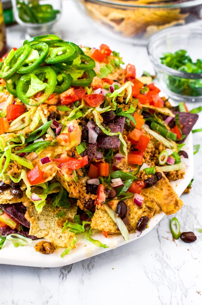 A close-up shot of a large plate of vegan nachos, piled high with walnut meat, dairy free nacho cheese sauce, and vegetables. Topped with sliced jalapenos.