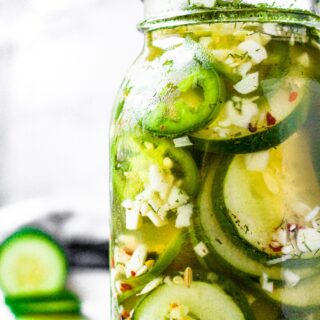 Close up head on shot of a large glass jar filled with pickled cucumber and jalapeno slices. There is a sliced cucumber in the background.