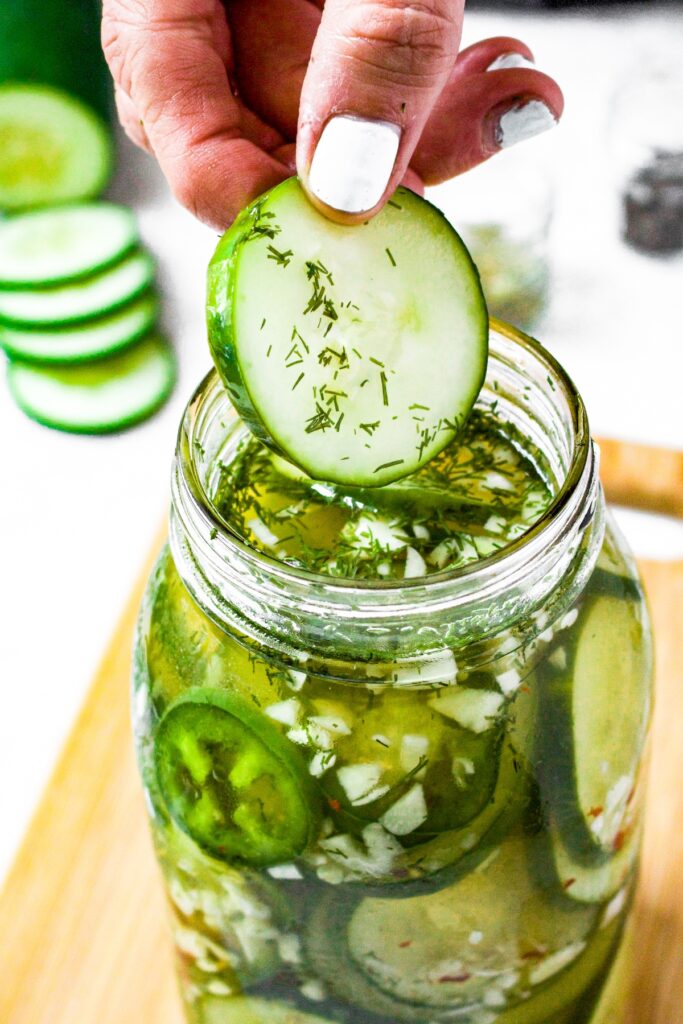 Overhead shot of a hand pulling a pickle slice out of a large mason jar. There are sliced cucumbers in the background.