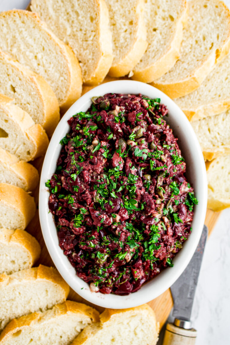 Overhead close up view of a white oval dish filled with vegan tapenade surrounded by baguette slices and topped with minced parsley