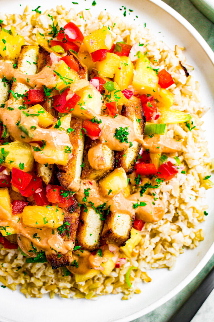 Overhead close up photo of half a round white plate topped with rice, pineapple tofu, pineapple jalapeno salsa, and drizzled with spicy vegan mayo