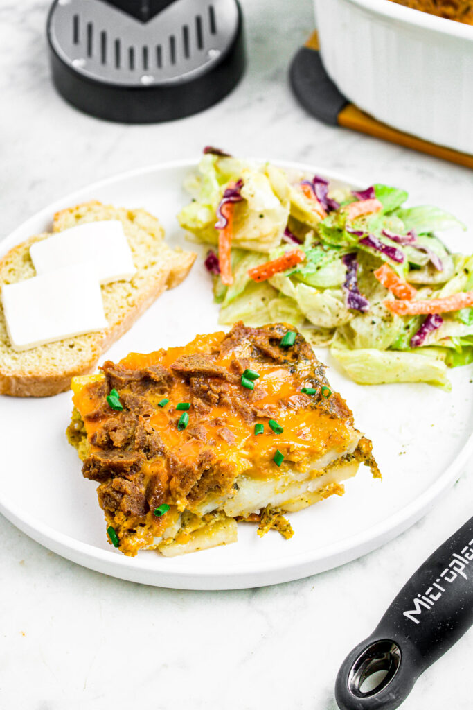 Close up head on photo of a slice of vegan potato casserole on a round white plate with salad and a slice of bread with butter pats