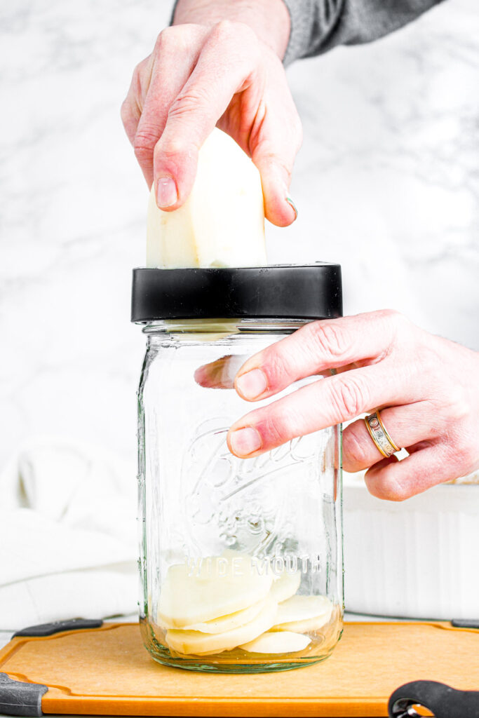 Head on photo of a hand running a potato across a jar top slicer as thinly sliced potatoes fall into the jar
