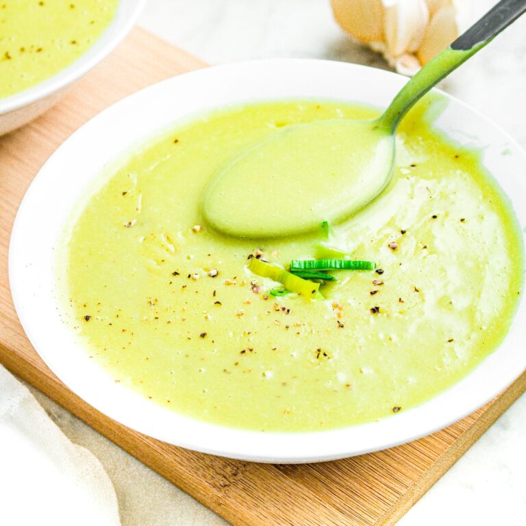 Close up overhead view of a round white bowl of dairy free leek and potato soup with a spoon sticking into it to show the creamy thick texture