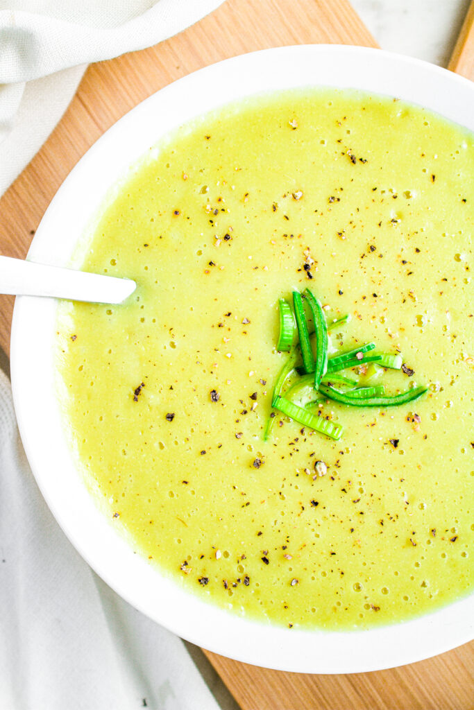 Overhead close up view of a round white bowl with vegan creamy potato leek soup topped with sliced leeks and black pepper