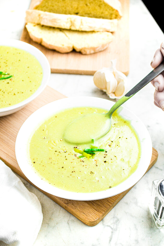 Overhead view of a round white bowl with creamy leek and potato soup with a spoon sticking into it and partially sliced loaf of bread behind it